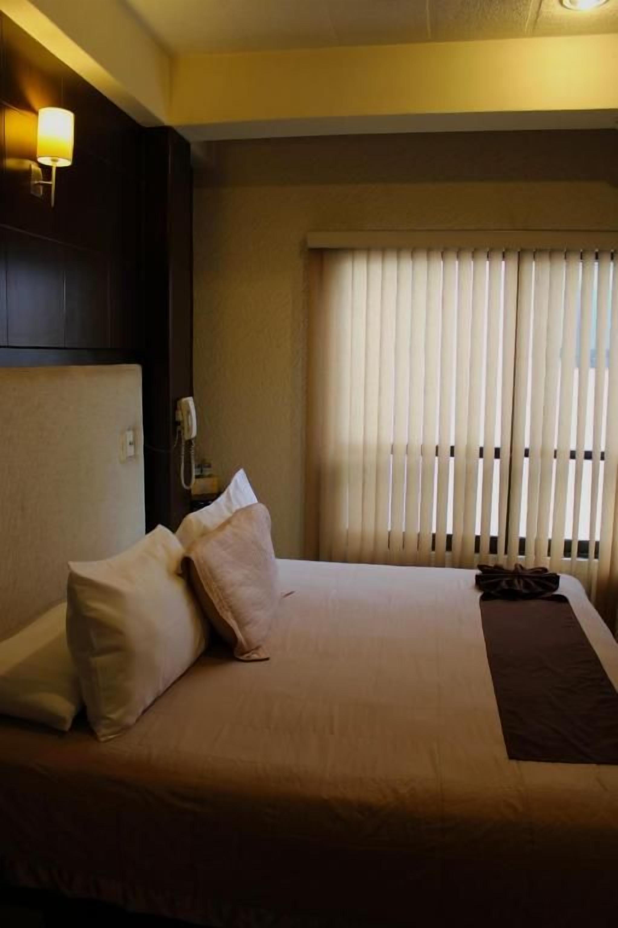 A hotel room with a bed, pillows, a nightstand, a lamp, and a window with blinds.