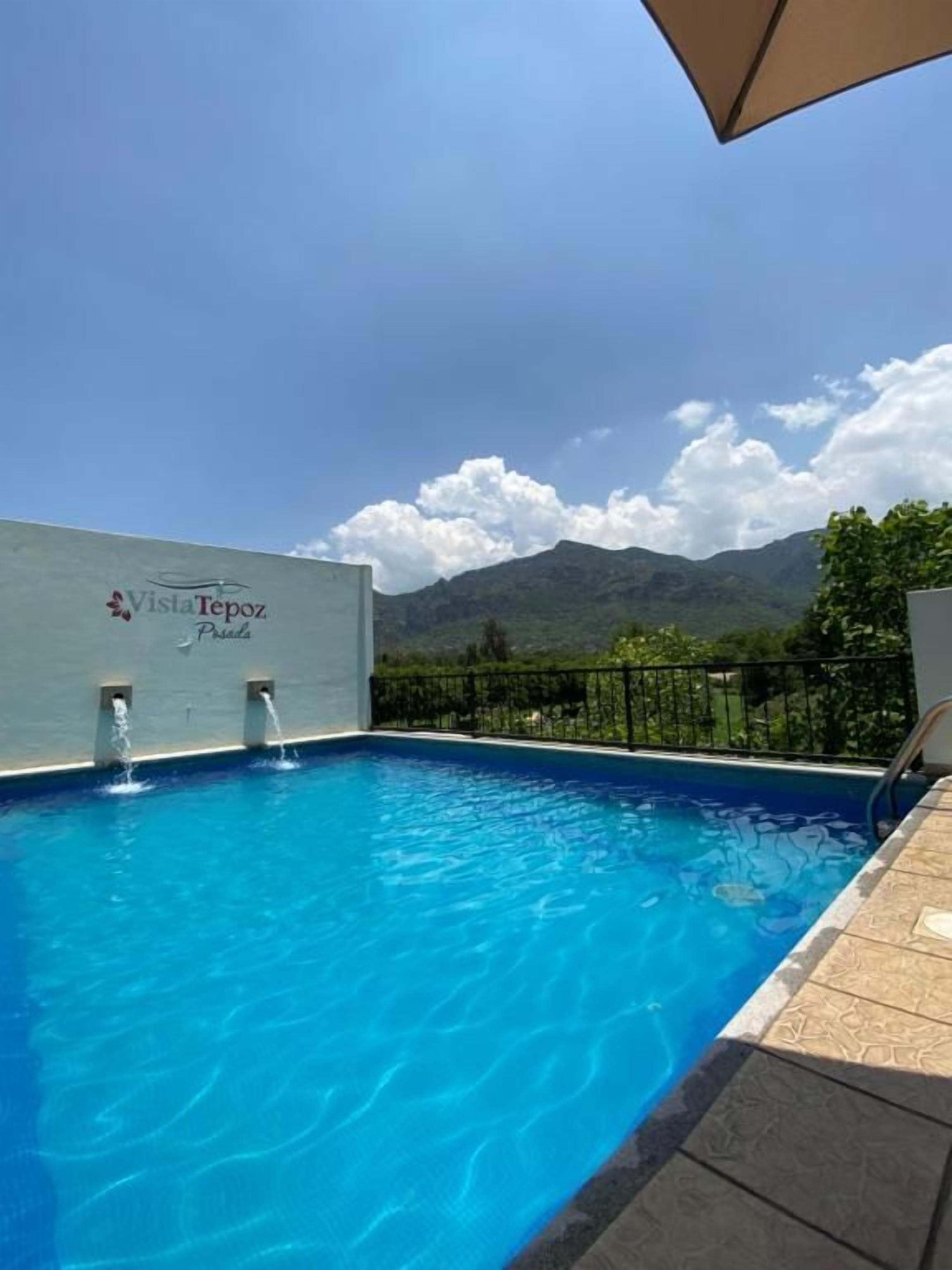 A swimming pool with a mountain view, a sign reading 'Vista Tepoz Beach,' and a clear sky.