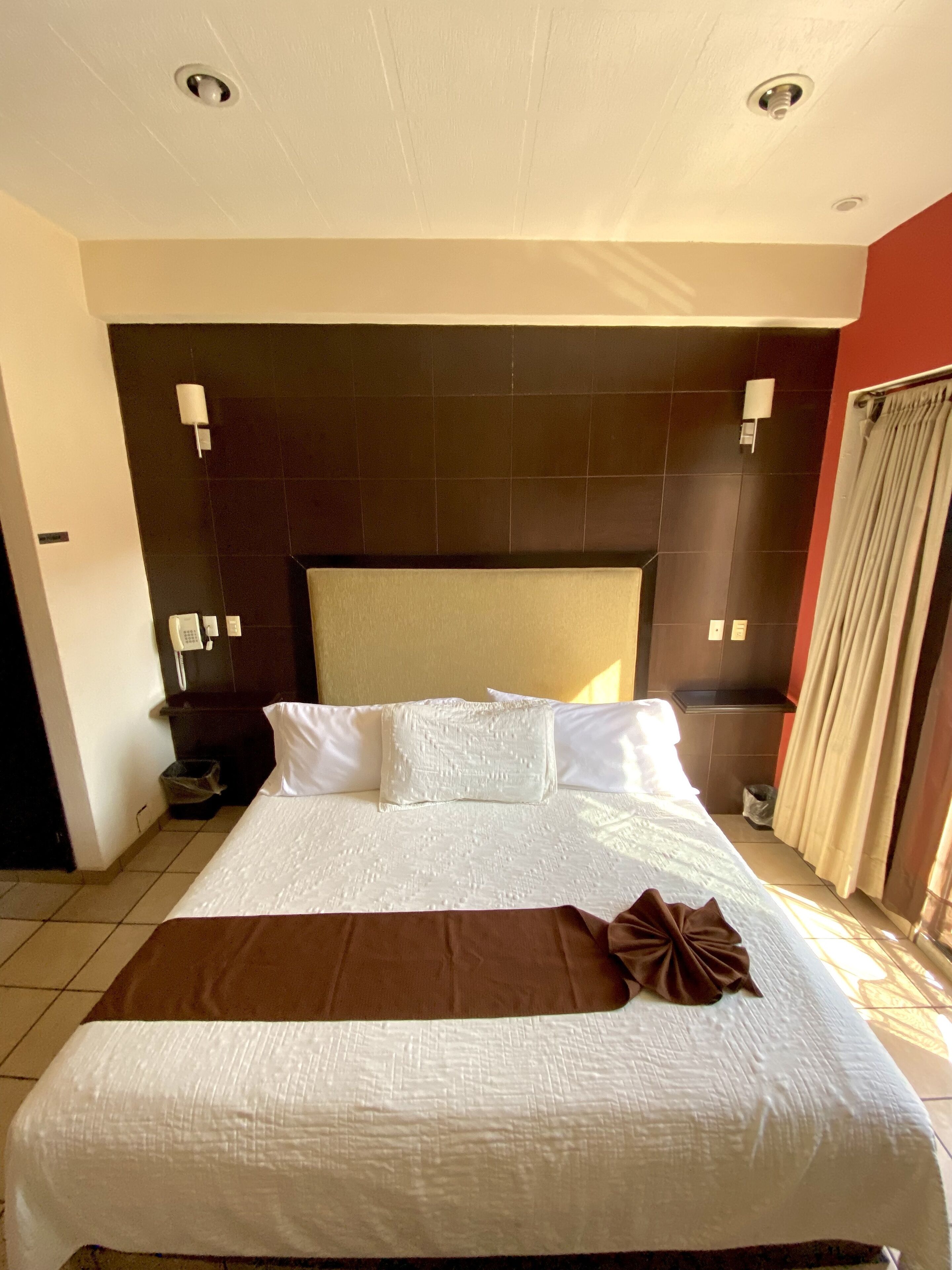 A hotel room with a large bed, a dark headboard, and a view of the outside through a window with curtains.
