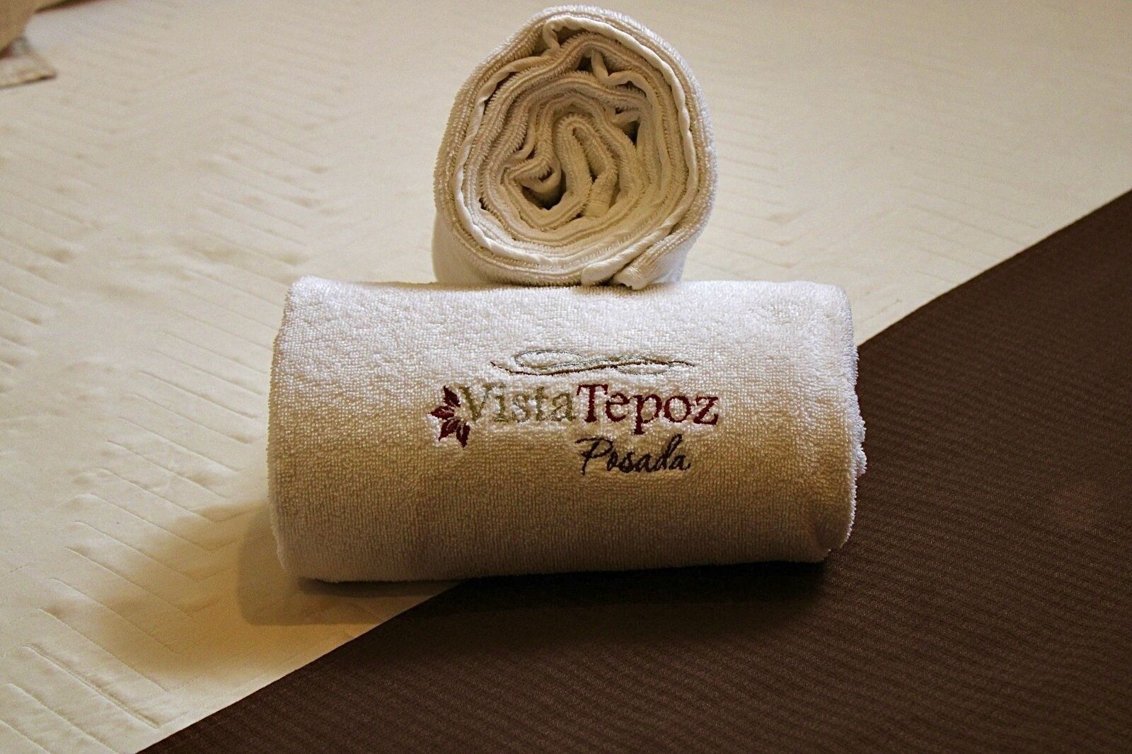 Ein ordentlich gerolltes weißes Handtuch mit aufgesticktem Text "Vista Tepoz Posada" auf einer strukturierten Oberfläche.