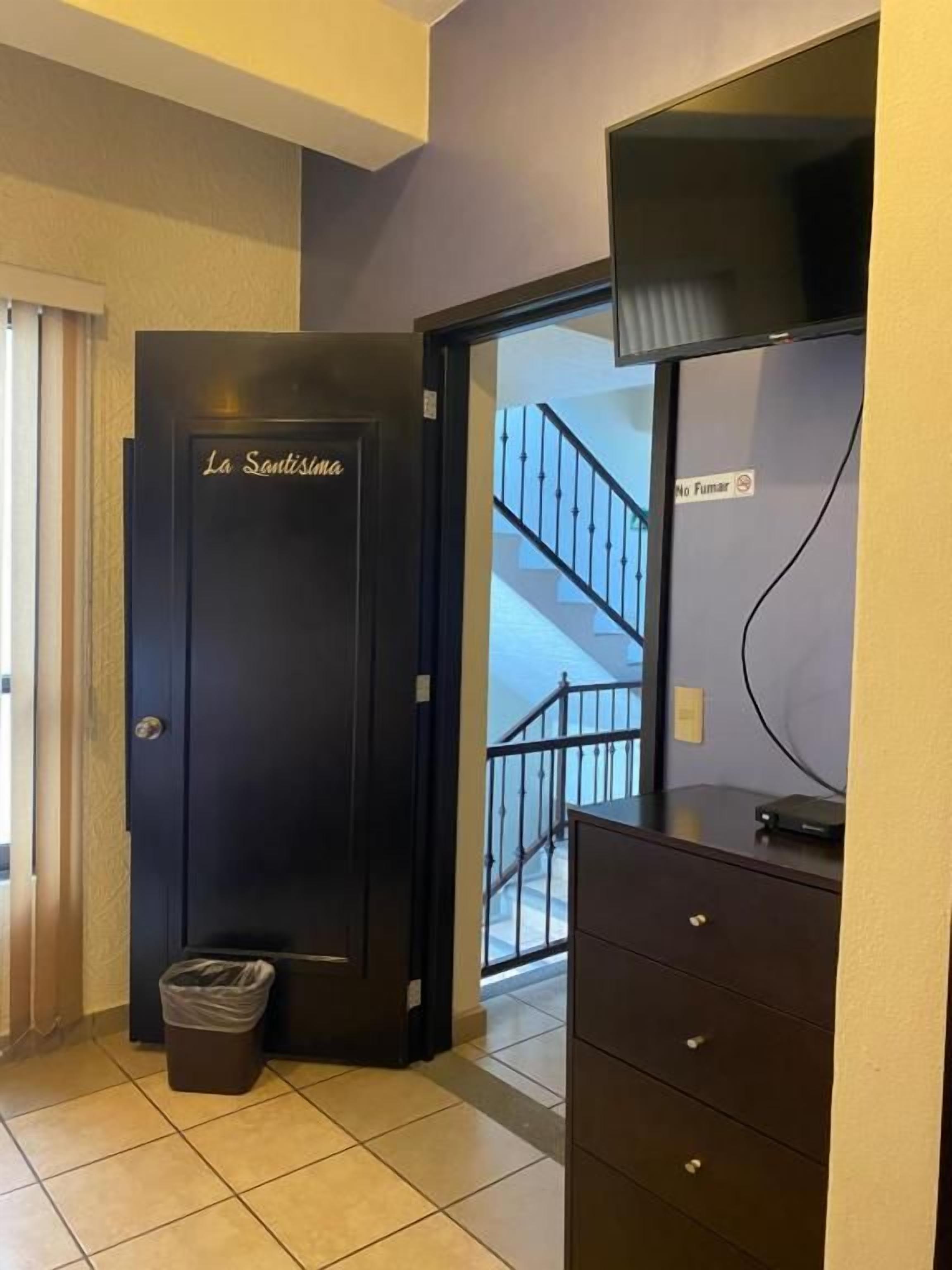 A hotel room with a door labeled 'La Santísima,' a trash bin, a dresser, and a staircase leading to another floor.