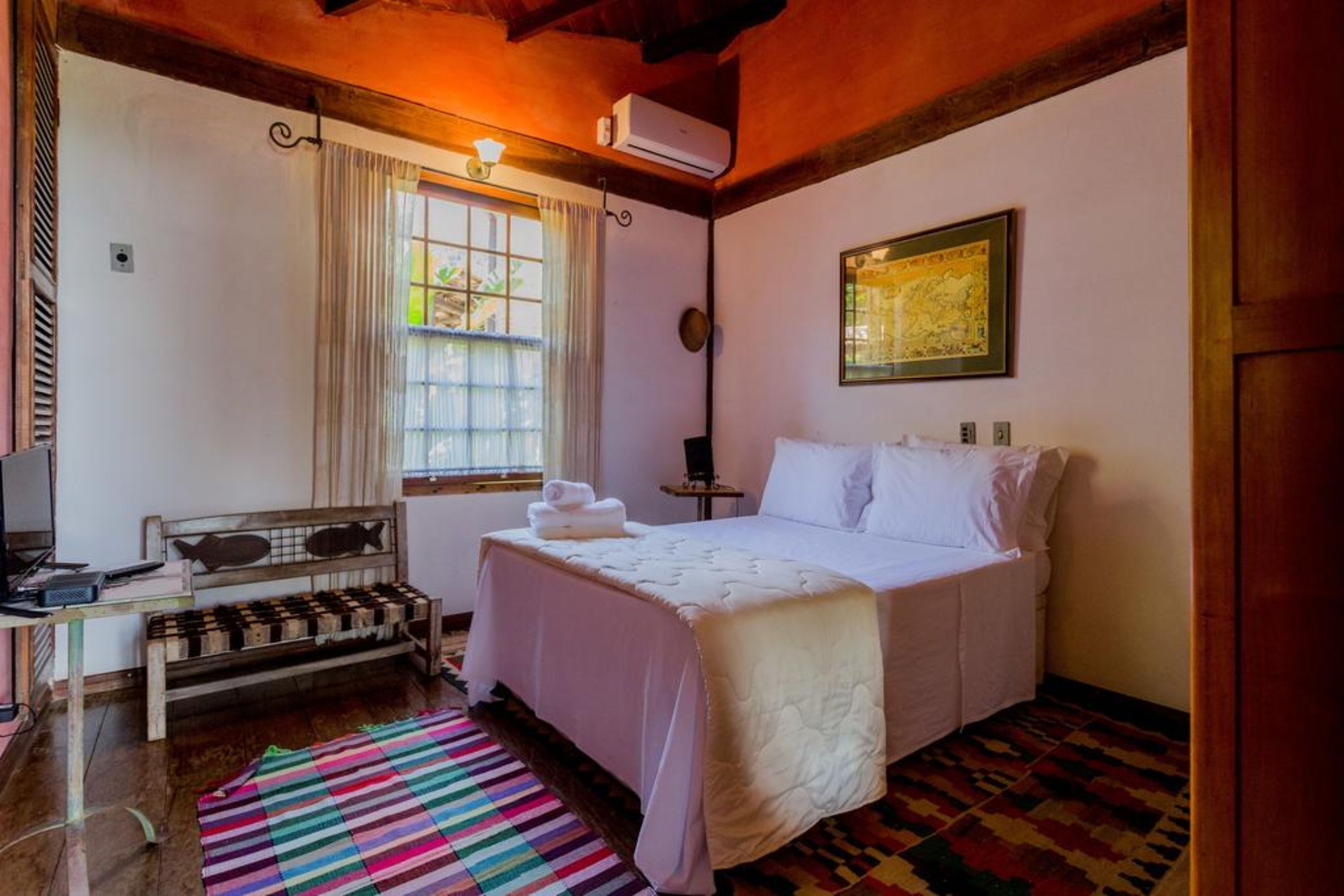 A bedroom with a bed, a desk, a chair, a window with curtains, and a wall-mounted air conditioner.