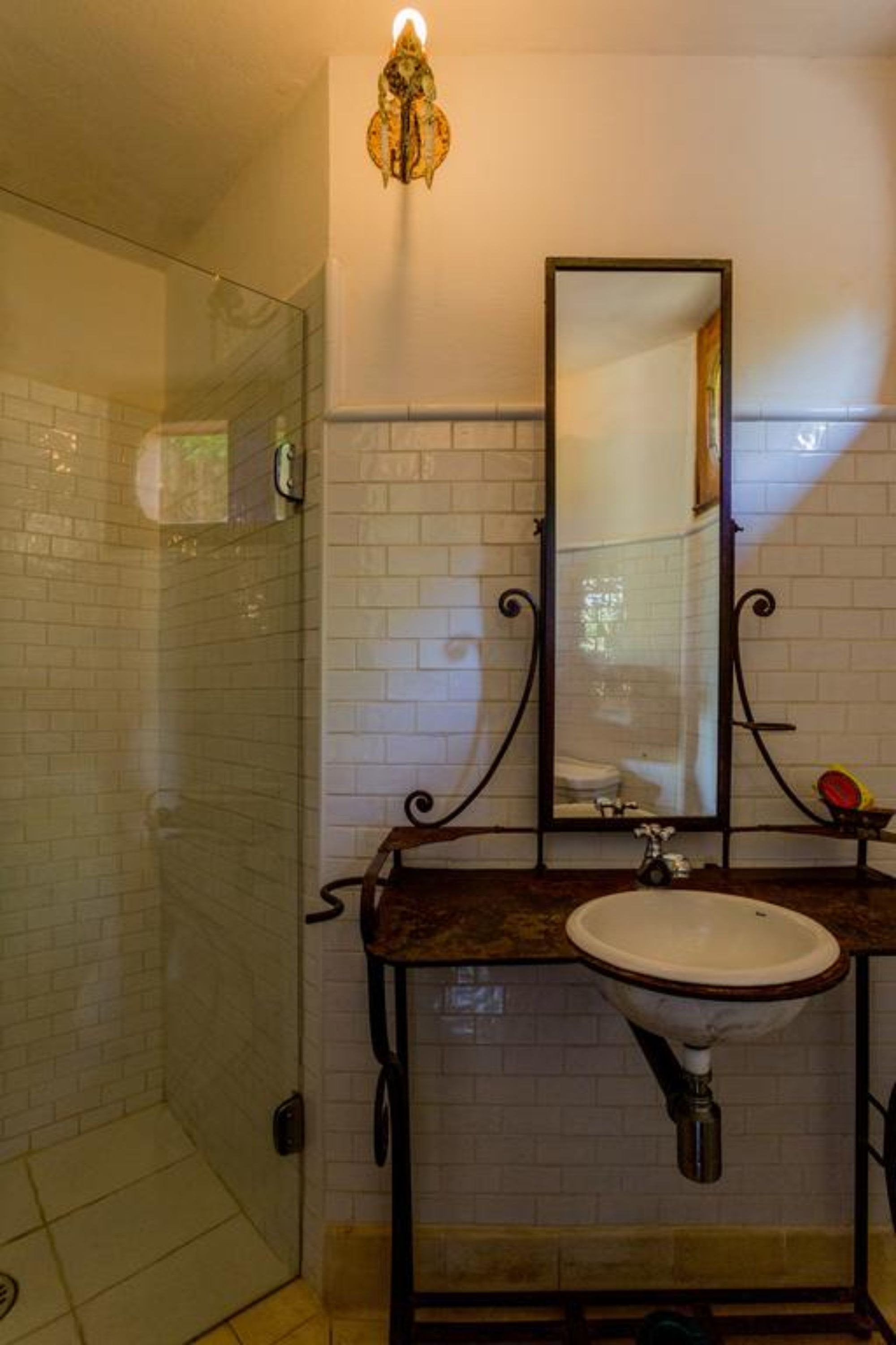 A bathroom with a glass shower enclosure, a white sink with a marble countertop, and a wall-mounted mirror with a decorative light fixture.
