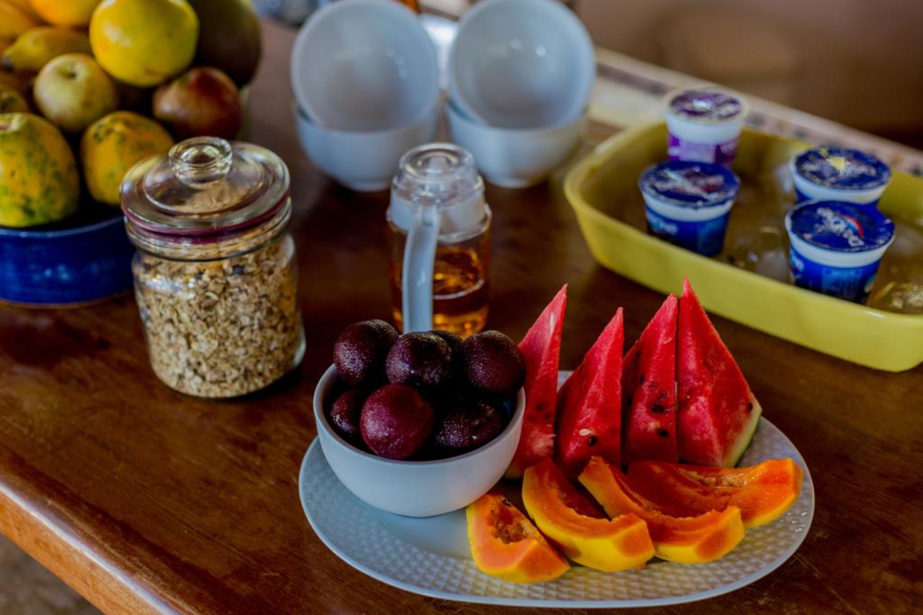 A wooden table with a bowl of cherries, sliced watermelon, and papaya. There are also jars of nuts and a container of honey.