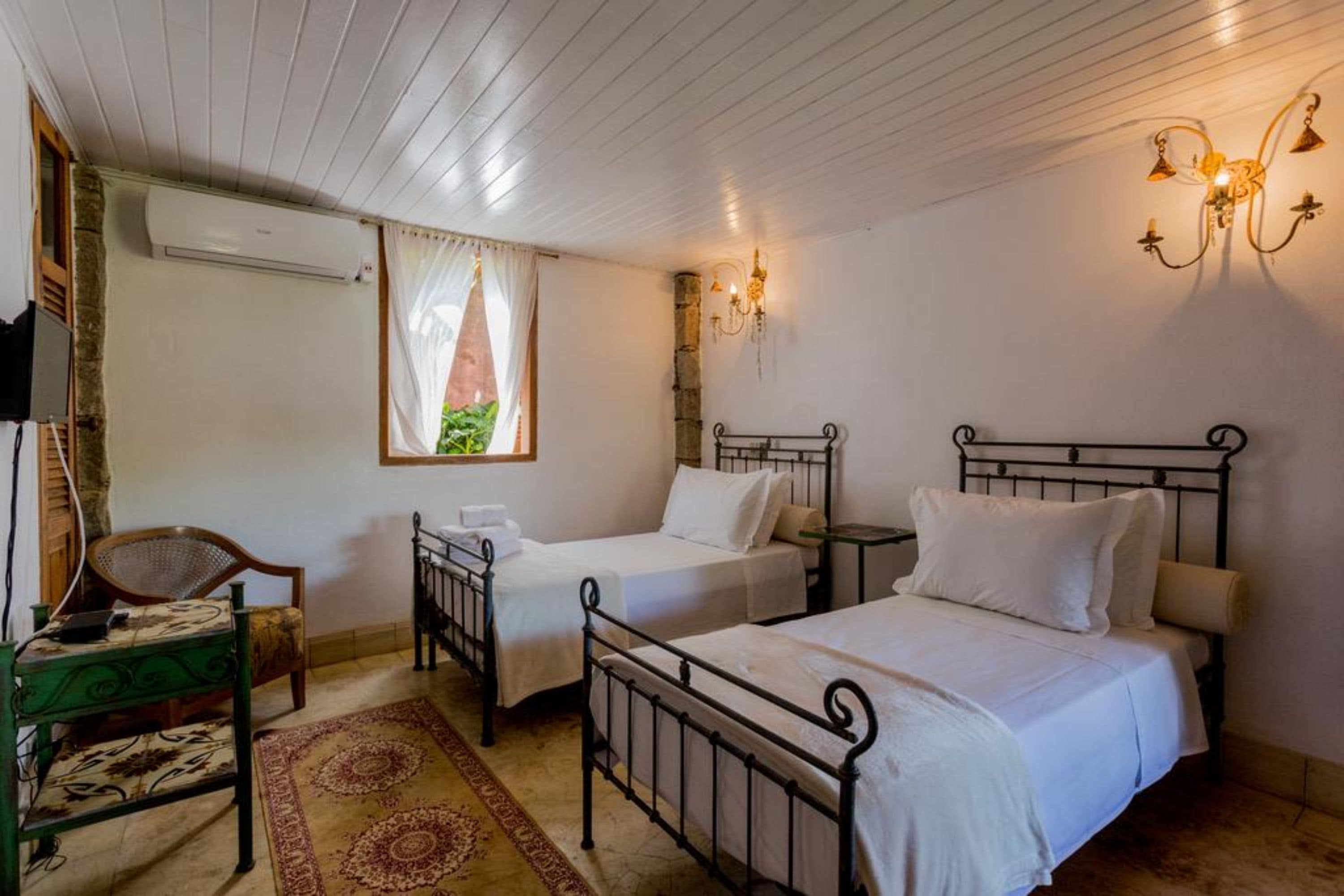 Quarto com duas camas de solteiro, uma cadeira, uma mesa pequena e uma janela com cortinas.