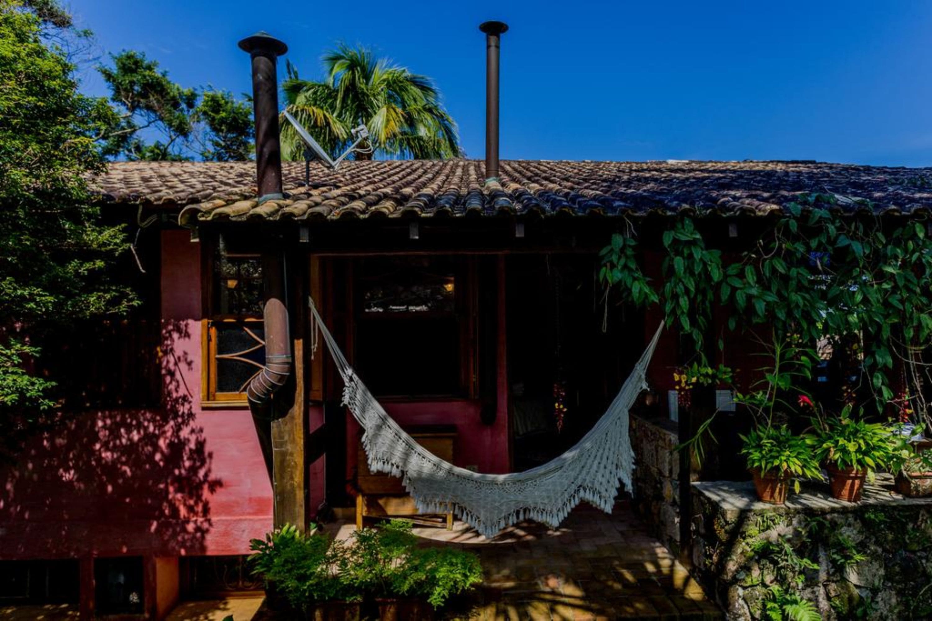 Uma casa rústica com telhado de telhas, chaminé e uma rede em frente.