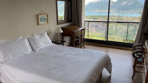 A bedroom with a large bed, a desk, a chair, a lamp, a framed picture on the wall, and a large window with a mountain view.