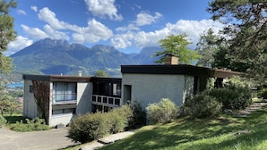 A modern two-story house with a balcony, surrounded by lush greenery and mountains in the background.