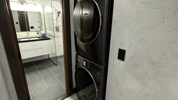 A laundry room with a washing machine and dryer, a glass door leading to a bathroom, and a wooden floor.