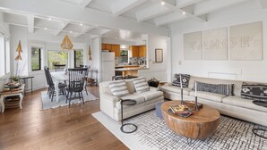 Uma sala de estar ampla com uma mesa de jantar, sofá e mesa de centro. É possível ver uma cozinha ao fundo, com armários de madeira e uma geladeira.