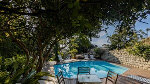 A wooden deck with a pool, surrounded by lush greenery and a stone wall. - Ilhabela Homestay (Ilhabela)