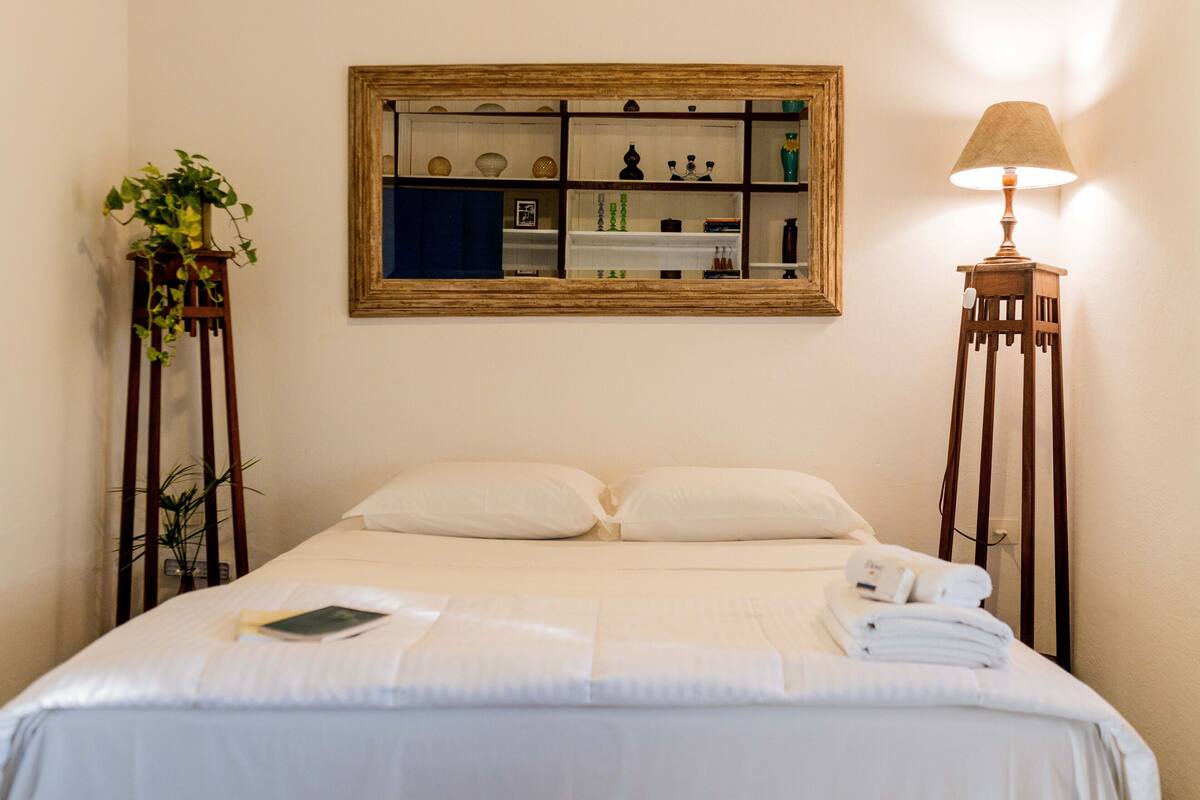Un letto ben rifatto con lenzuola bianche, una lampada da comodino e un piccolo tavolino con un libro e un telecomando.