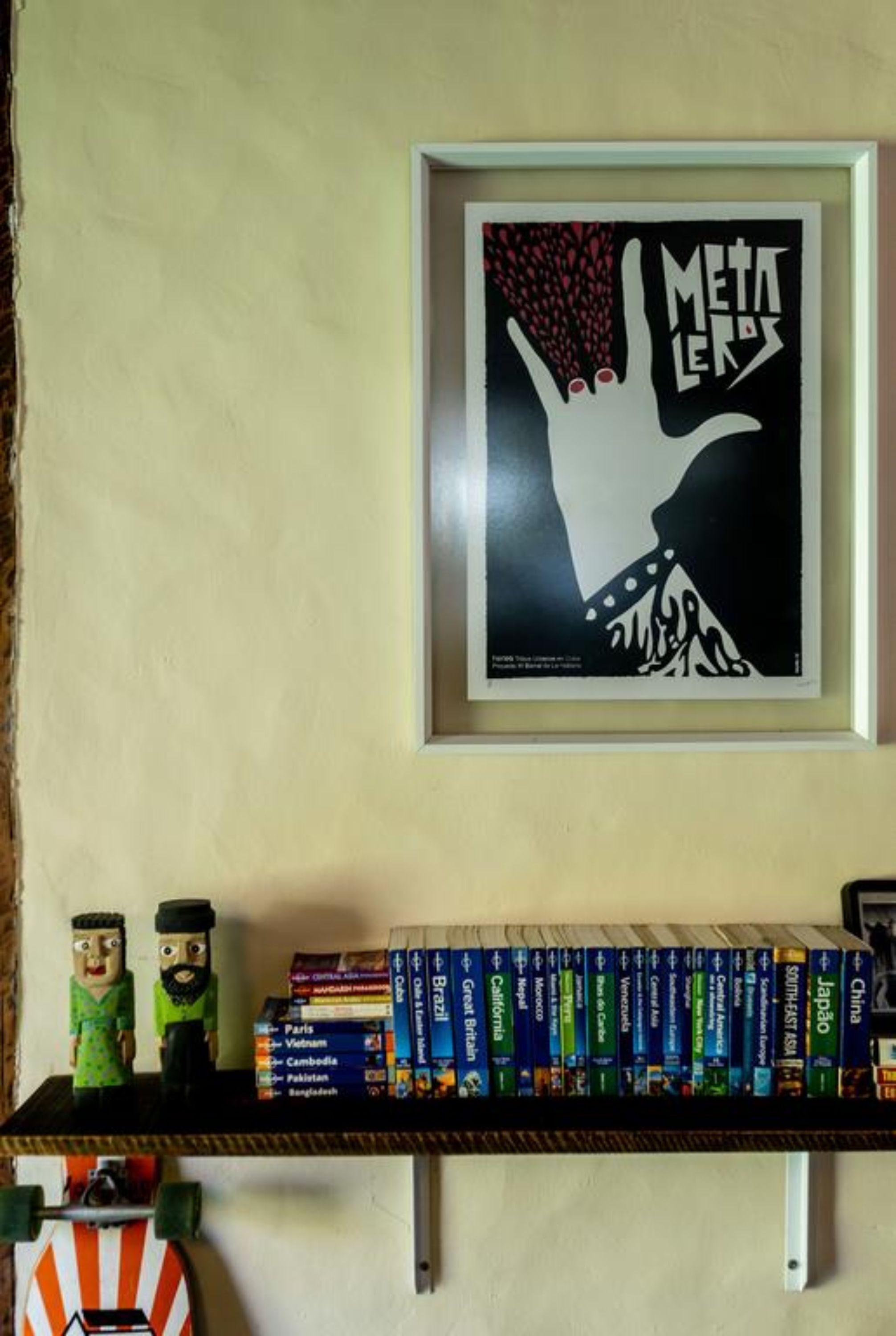Um cartaz emoldurado de uma mão com o texto "METAL LEVER" em uma parede. Abaixo, uma estante com livros de viagens pode ser vista.
