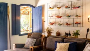 A cozy living room with a wooden ceiling, a large window with blue shutters, a wall-mounted sailboat artwork, and a comfortable sofa set. - Ilhabela Homestay (Ilhabela)