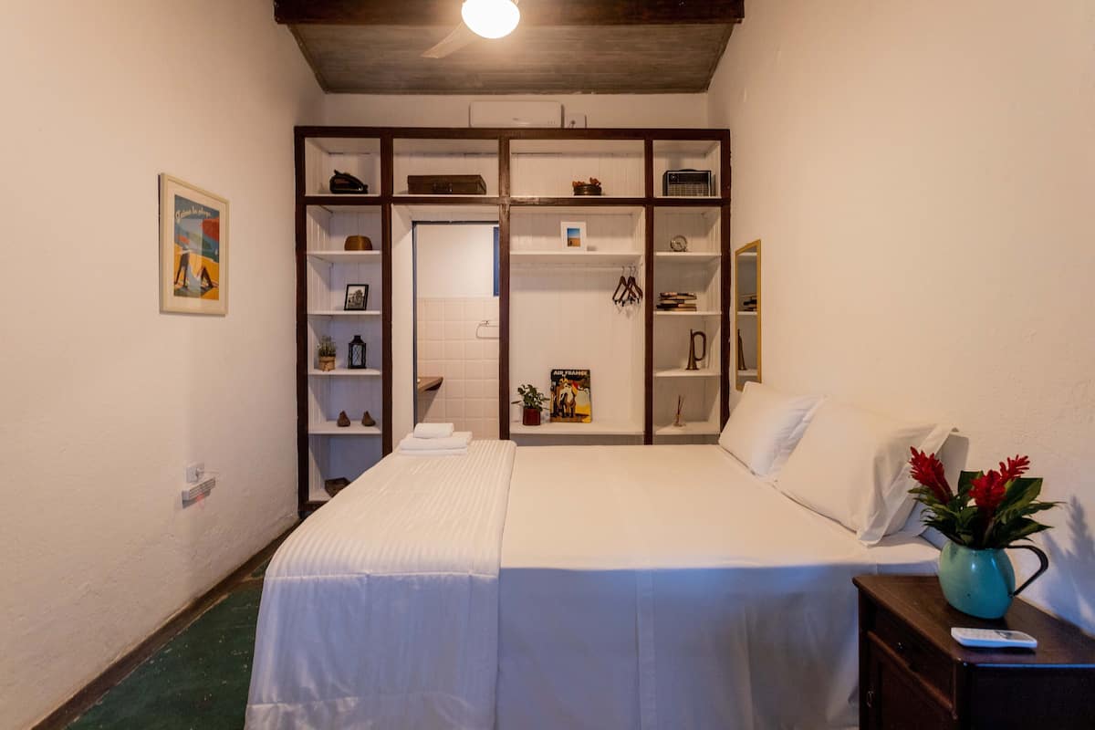 Una camera da letto con un letto, un comodino, una mensola con dei libri, un quadro incorniciato e una porta che dà su un'altra stanza.