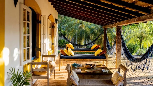 A hammock chair with cushions, a wooden bench, and a table with a bowl and a book. - Ilhabela Homestay (Ilhabela)