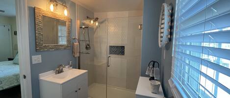 Un baño moderno con cabina de ducha de vidrio, tocador con espejo y ventana con persianas.