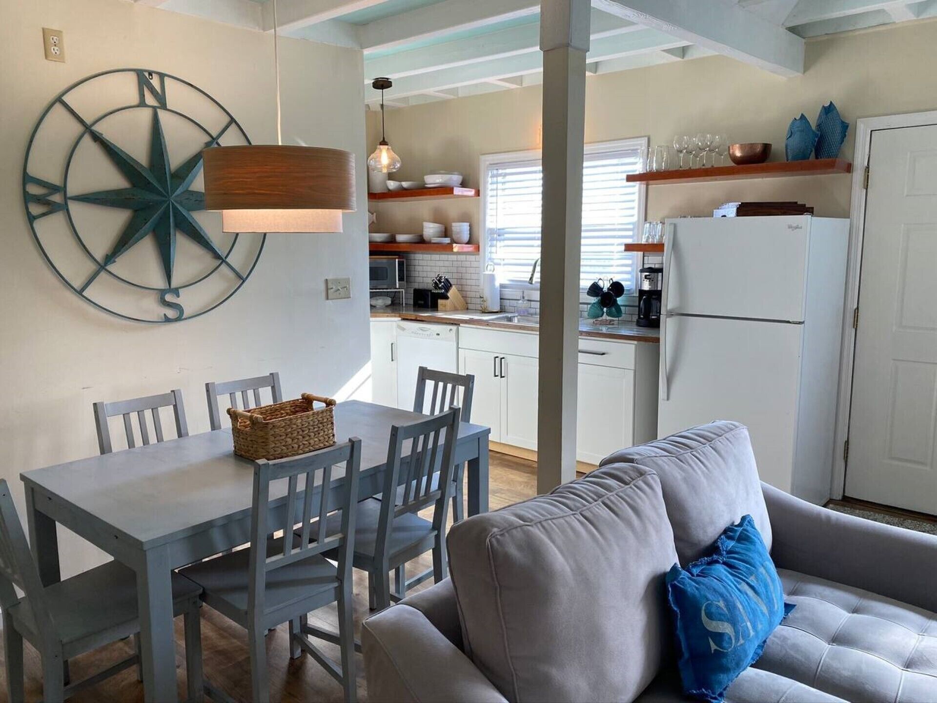 Una cocina moderna con armarios blancos, una mesa de comedor con sillas, un adorno decorativo grande con forma de brújula y una sala de estar acogedora con sofá y cojines.