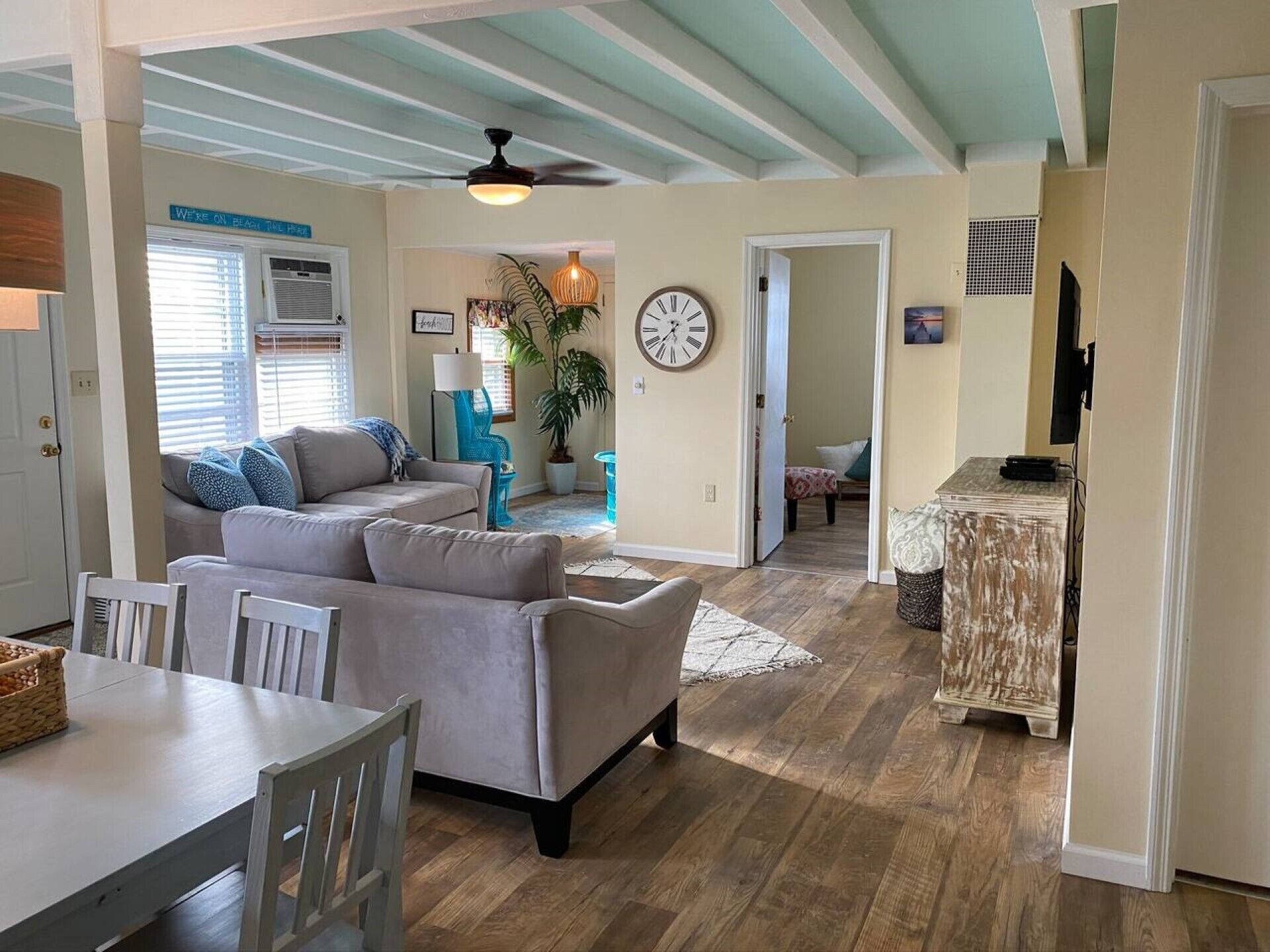 A spacious living area with a grey sofa, a dining table with chairs, a ceiling fan, and a clock on the wall.