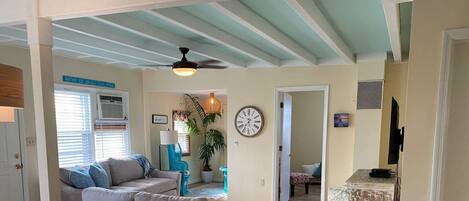 A spacious living area with a grey sofa, a dining table with chairs, a ceiling fan, and a clock on the wall.