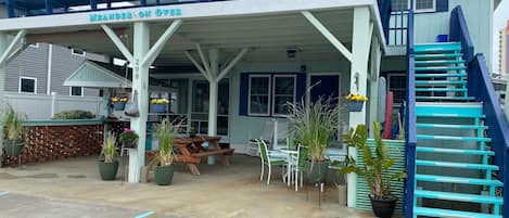 A covered outdoor seating area with a picnic table and chairs, a blue staircase, and a sign reading 'MEANDER ON OVER'.