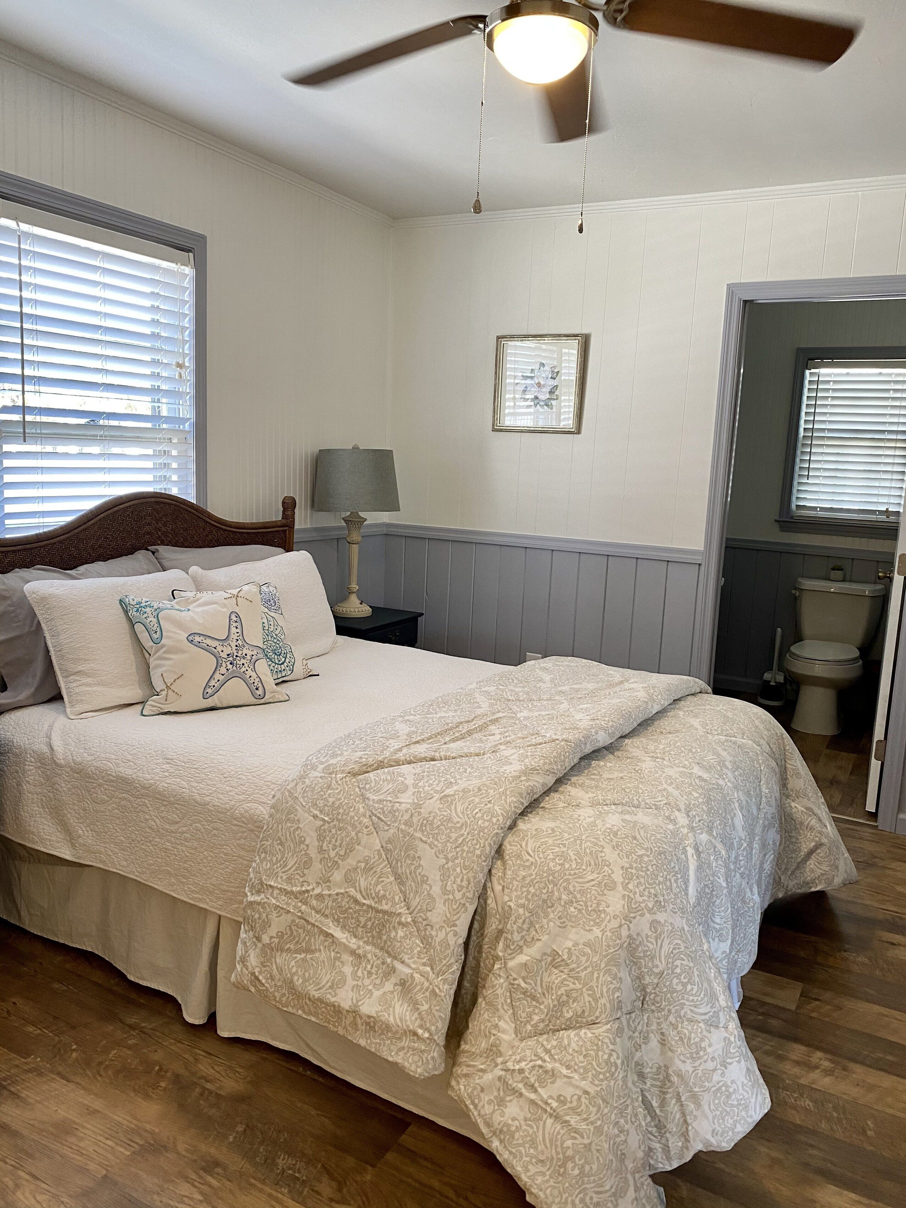 A bedroom with a bed, a ceiling fan, a nightstand with a lamp, and a window with blinds.