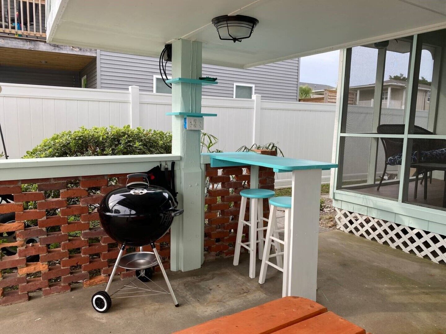 Un patio exterior con una parrilla, una mesa con taburetes y una ventana con vista a una zona residencial.