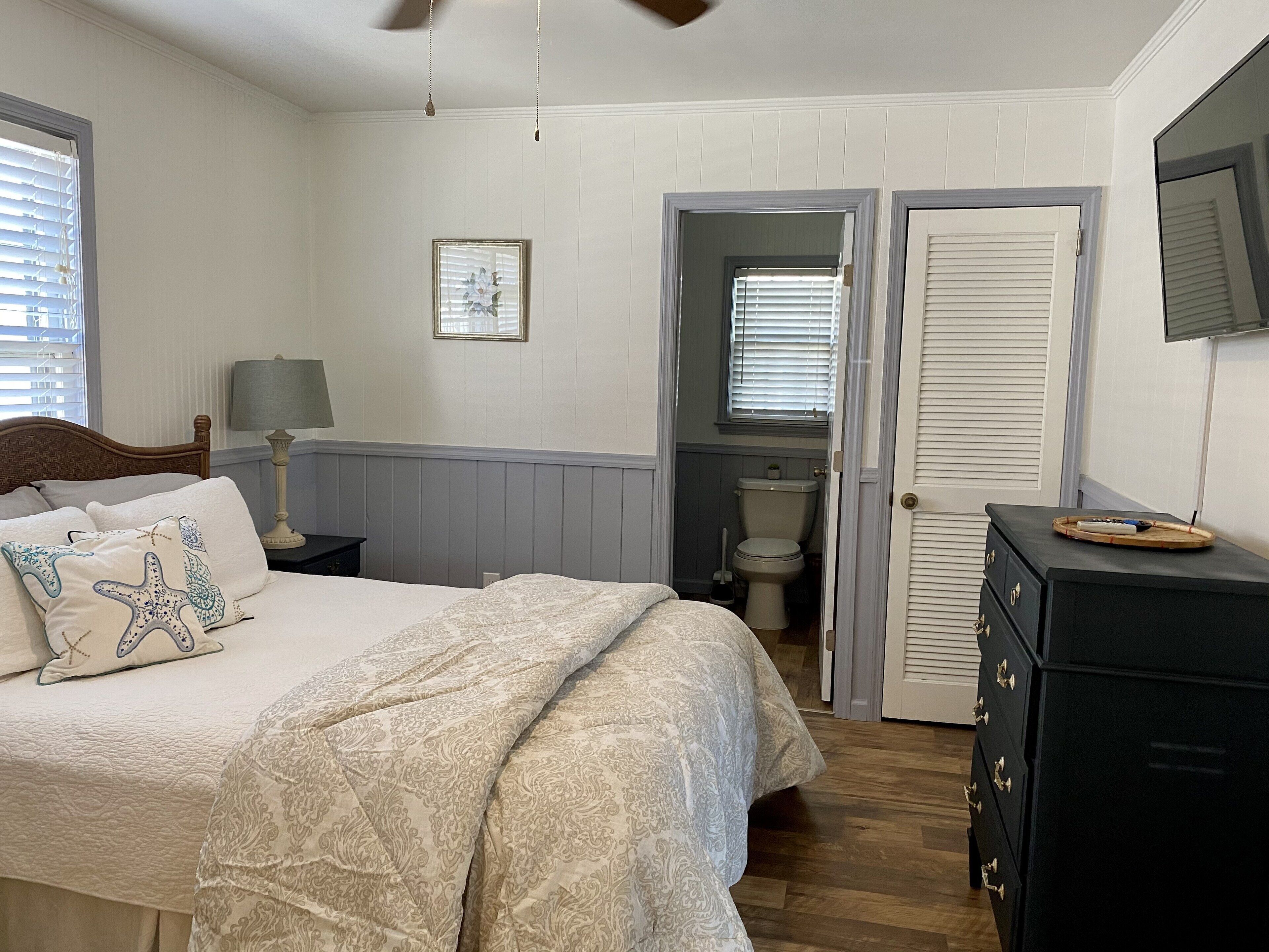 A bedroom with a bed, bedside table, lamp, dresser, and a door leading to a bathroom.