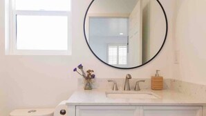 Une salle de bain avec un lavabo blanc, un miroir rond, des toilettes et un petit vase avec des fleurs.