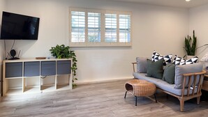 Un salon avec un téléviseur, une fenêtre avec des stores, un canapé avec des coussins, une plante et un plancher en bois.