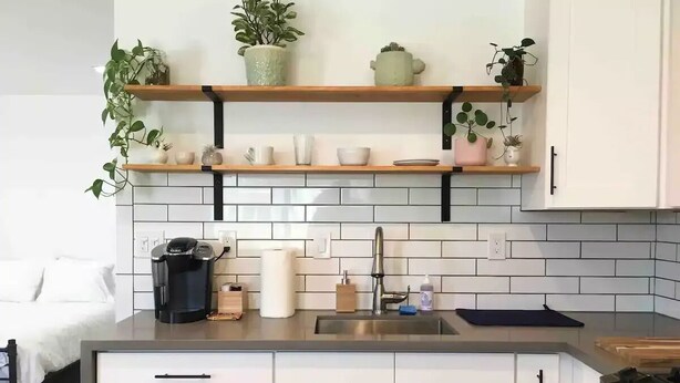 Une cuisine moderne avec des meubles blancs, un plan de travail noir et un pare-feu en carrelage. Des étagères au-dessus de l’évier servent à ranger des plantes et des ustensiles de cuisine.