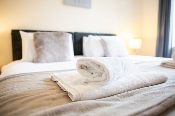 Een net opgemaakt bed met een opgerolde witte handdoek op de voorgrond, een hoofdbord en een lamp op het nachtkastje.