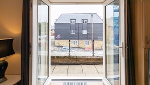 Una habitación con puerta corrediza de vidrio que da a un balcón. El balcón tiene baranda metálica y vista a una zona residencial con casas y coches aparcados.