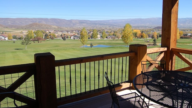 Una terraza de madera con una mesa y sillas, con vista a un campo verde con un estanque y casas a lo lejos.