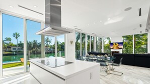 Una cocina moderna con una isla grande, un comedor con mesas de vidrio y vistas a una piscina y palmeras.