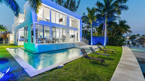 Una casa moderna de dos pisos con piscina, mobiliario exterior y palmeras.