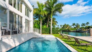 Una piscina con sillones de descanso, un edificio blanco con amplias ventanas y palmeras al fondo.