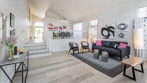 Uma sala de estar espaçosa com uma escada que leva a um andar superior, um sofá cinza escuro com almofadas rosa e uma mesa de centro de madeira.