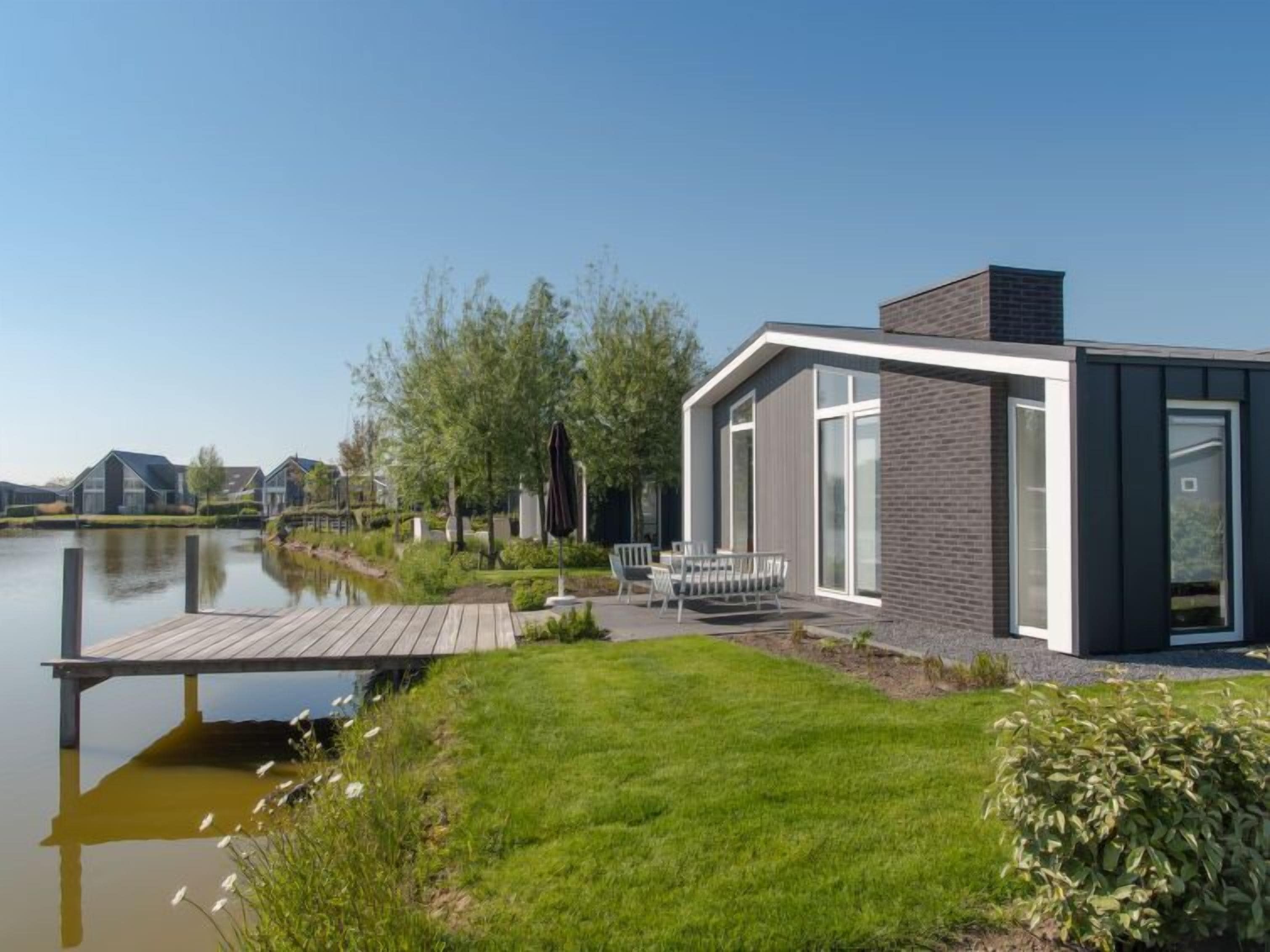 Ein modernes Haus mit einer Holzsteganlage, die sich in einen ruhigen See erstreckt, umgeben von viel Grün und einem klaren Himmel.
