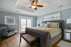 A bedroom with a bed, a chair, a bench, a ceiling fan, and artwork on the walls. - Luxurious Sunset Beach Home On The Water With Pool, Boat Dock and Golf Cart (Sunset Beach)