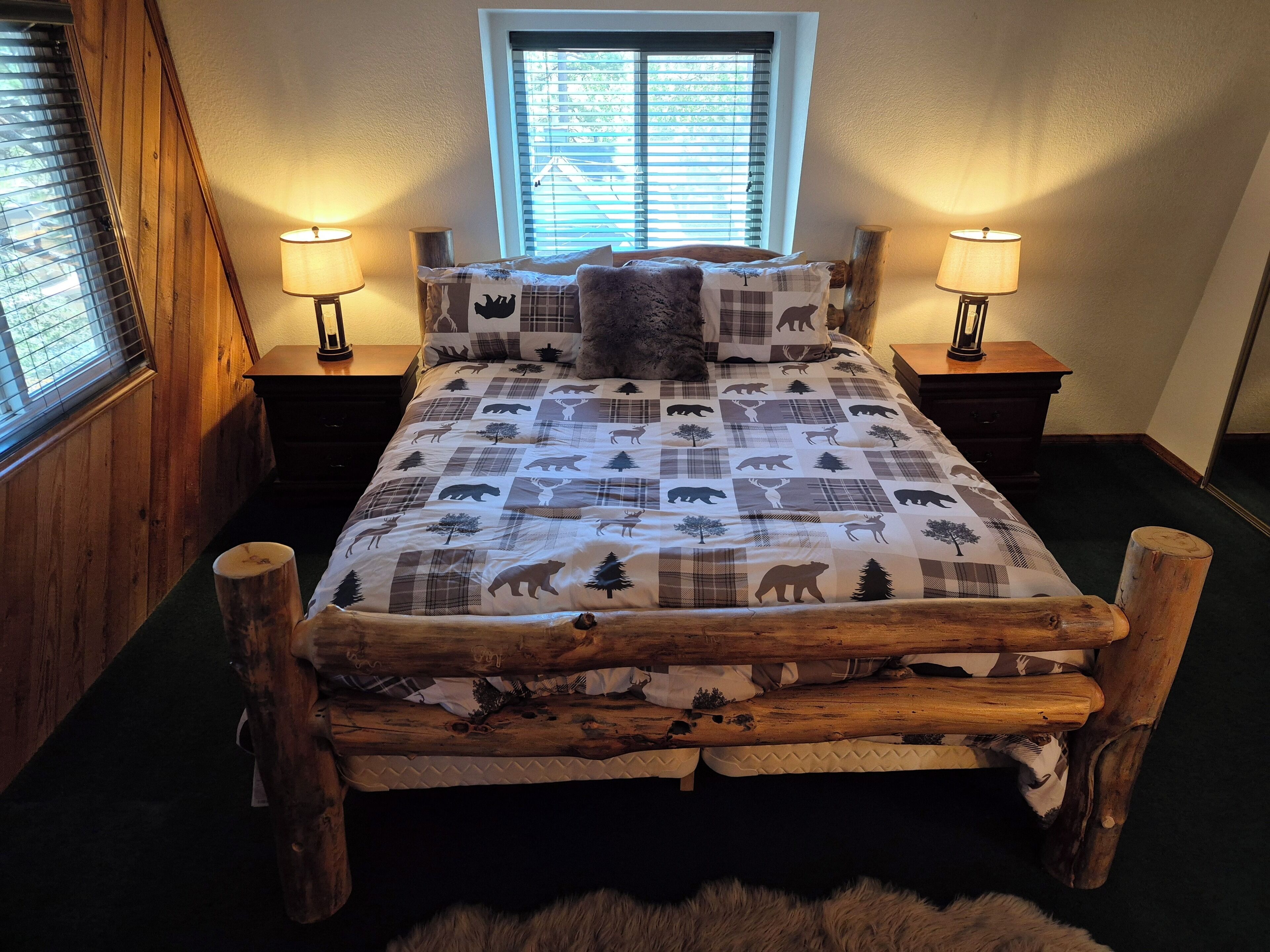 Una cama con estructura de madera y ropa de cama a cuadros con estampados de animales. Hay dos mesitas de noche con lámparas, una ventana con persianas y un cabecero de madera.