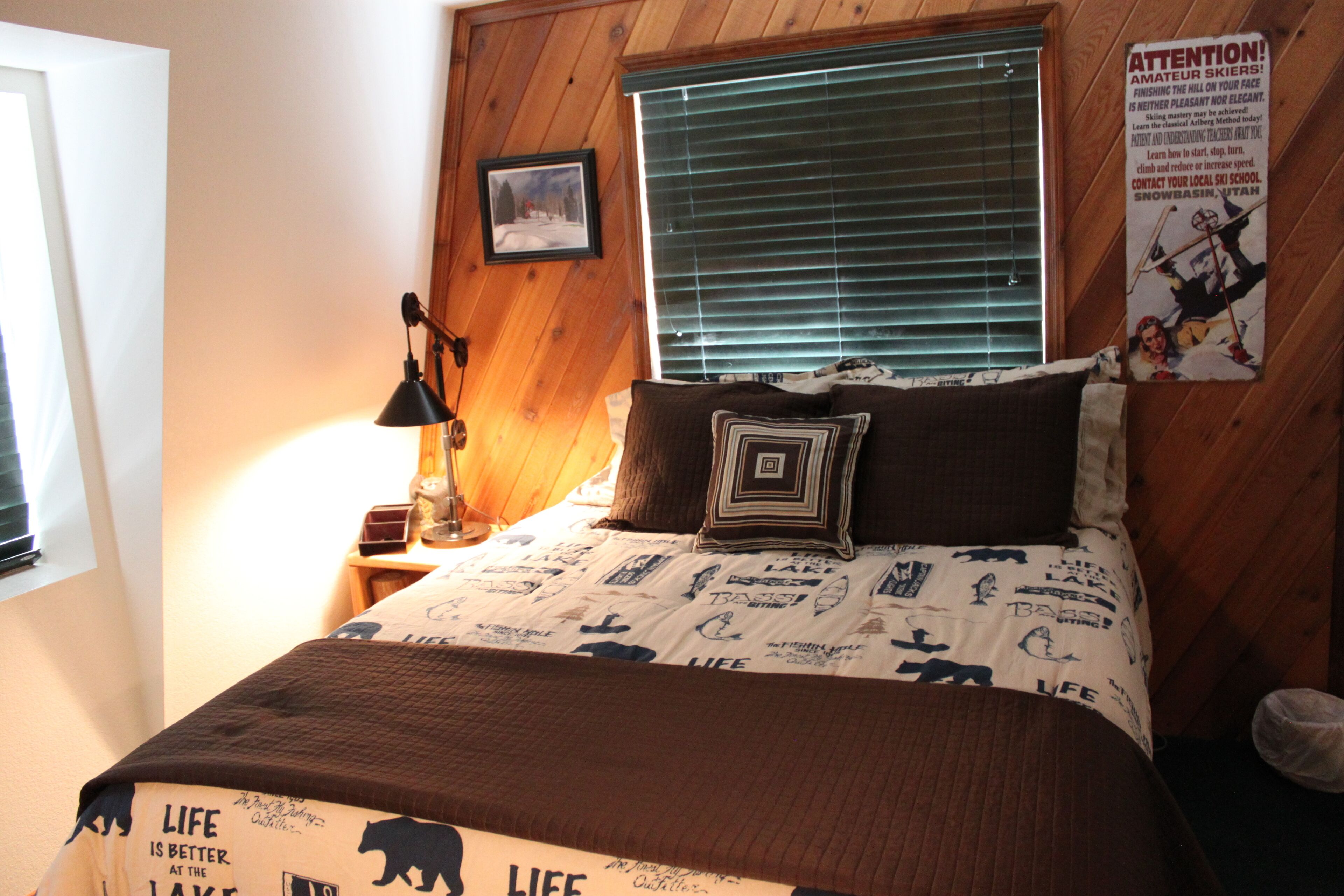 Un dormitorio con cabecera de madera, cama con ropa de cama estampada, lámpara de noche, un cuadro enmarcado en la pared y un cartel en la pared.
