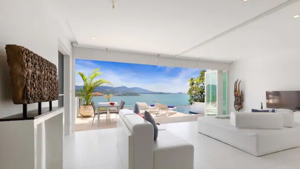 A modern living room with a large window overlooking a scenic view of a body of water and mountains. There is a white sofa, a dining table with chairs, and a TV on a stand. - Luxury Apartment Unique Residences (Koh Samui)