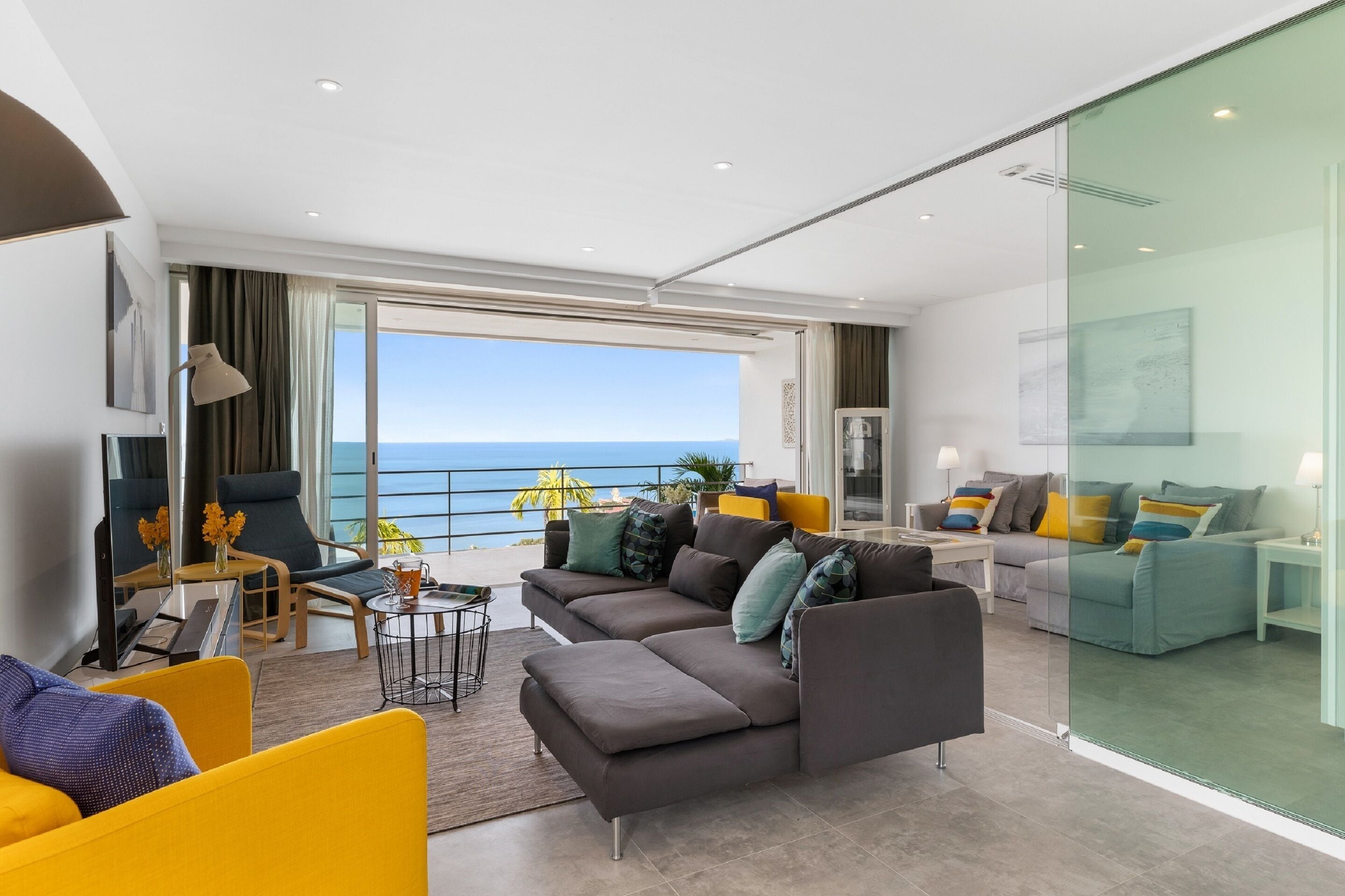 A modern living room with a large sectional sofa, a yellow armchair, and a glass partition wall.