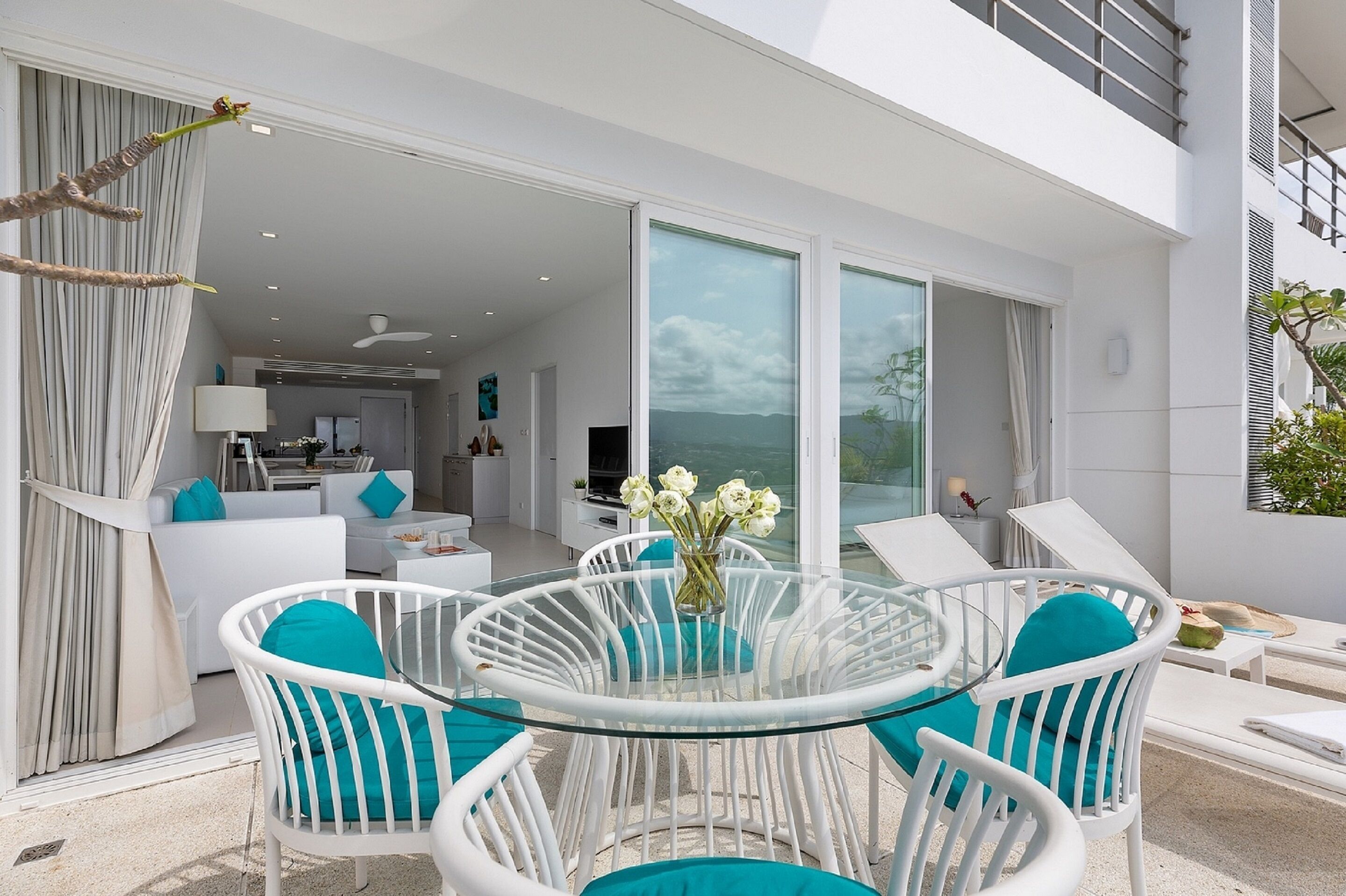 A modern outdoor dining area with a glass table and white chairs, a kitchen in the background, and a view of the sea.
