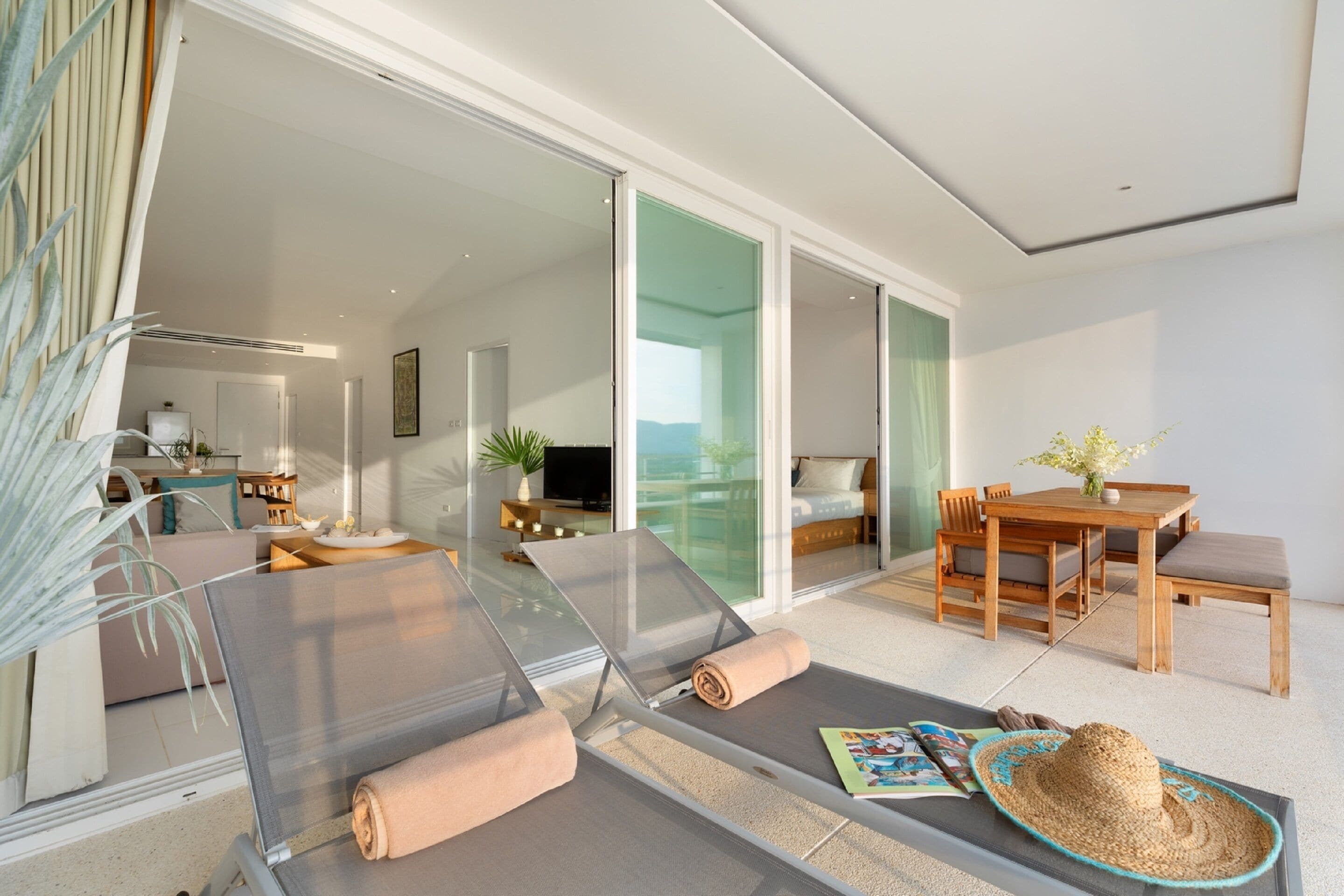 A modern hotel room with a glass sliding door leading to a balcony, a wooden dining table, and a lounge area with a sun lounger.
