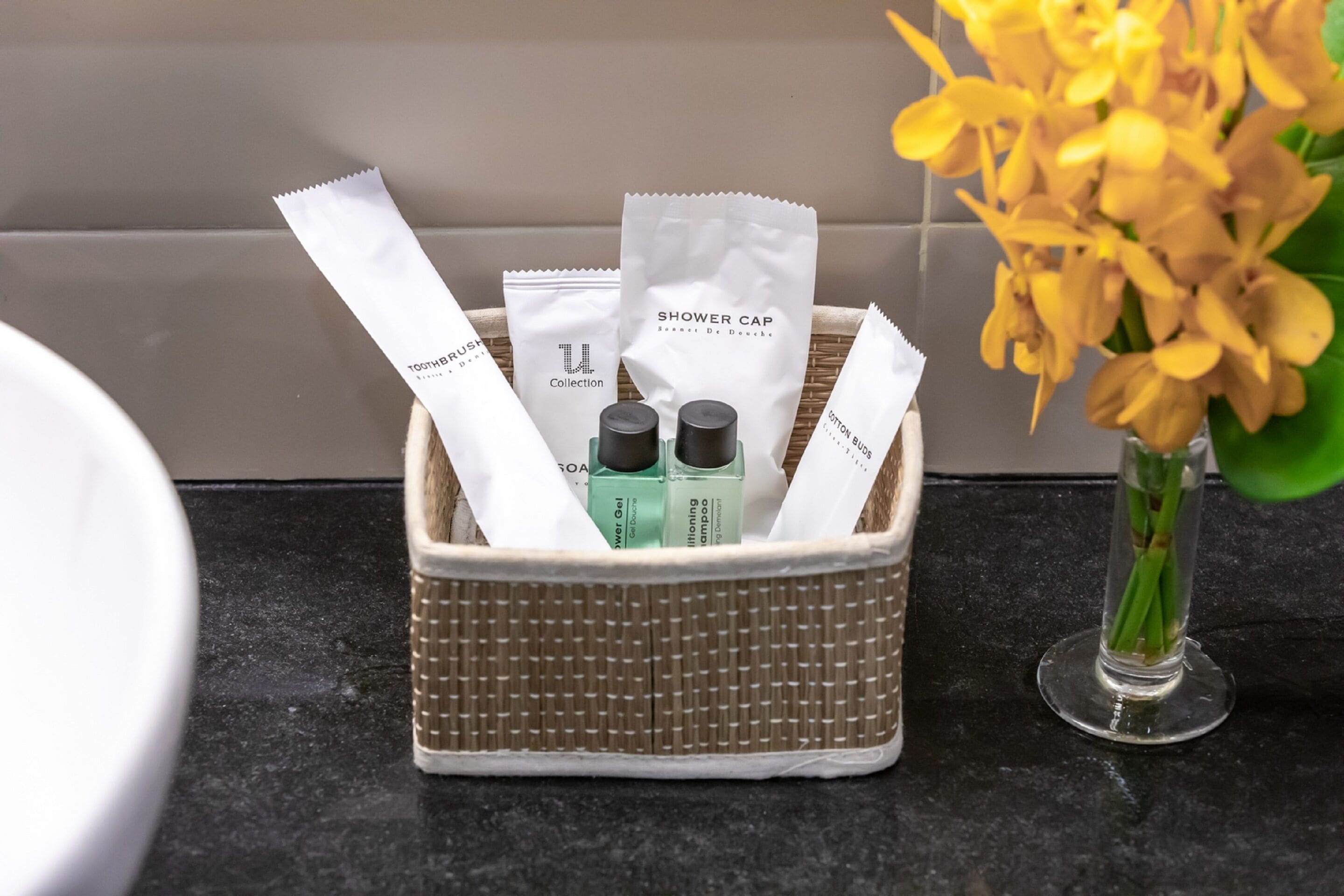 A bathroom sink area with toiletries in a basket, a shower cap, and a vase with flowers.