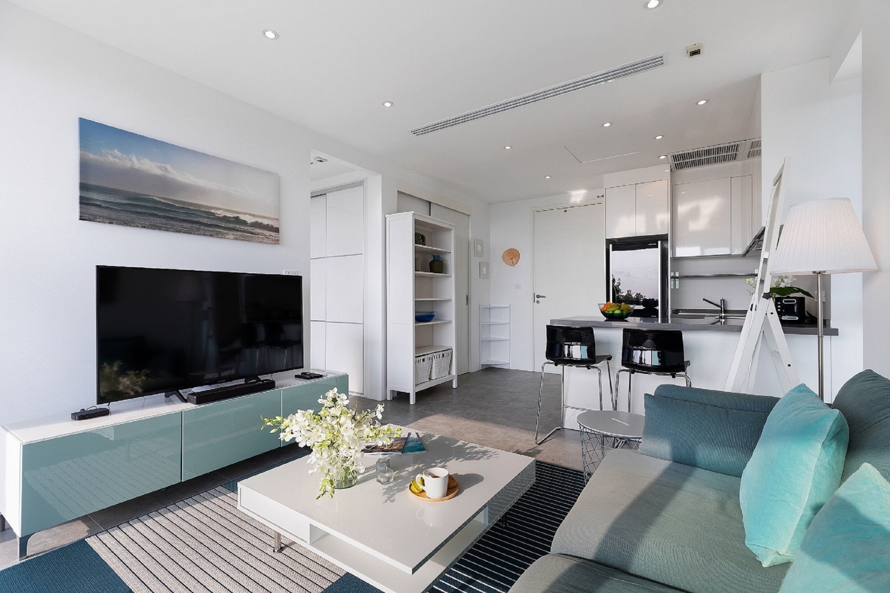 A modern living room with a flat-screen TV, a sofa, a coffee table with flowers, and a kitchen area with a fridge, microwave, and cabinets.