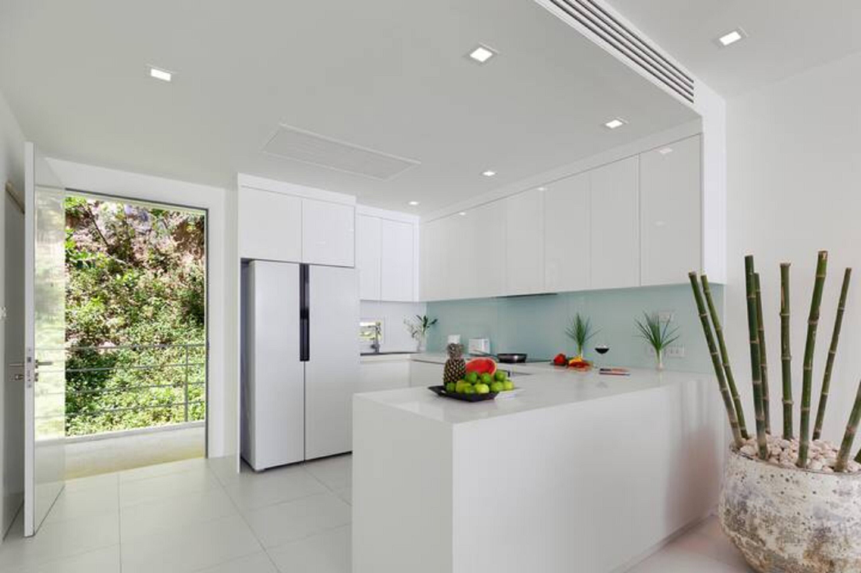 A modern kitchen with white cabinetry, a central island with fruit, and a view of greenery outside.