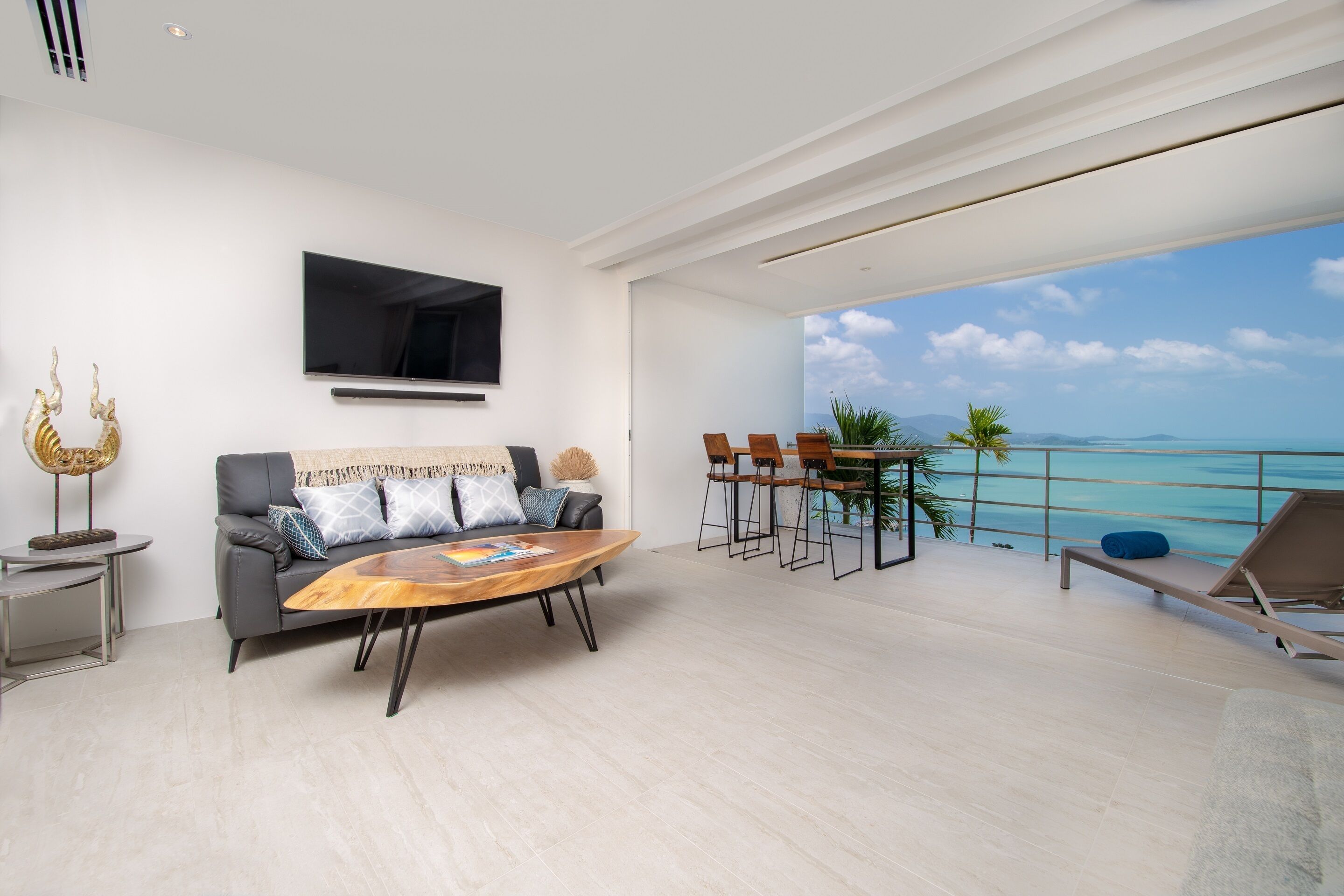 A modern living room with a sofa, coffee table, and a TV. There's a balcony with a view of the ocean.